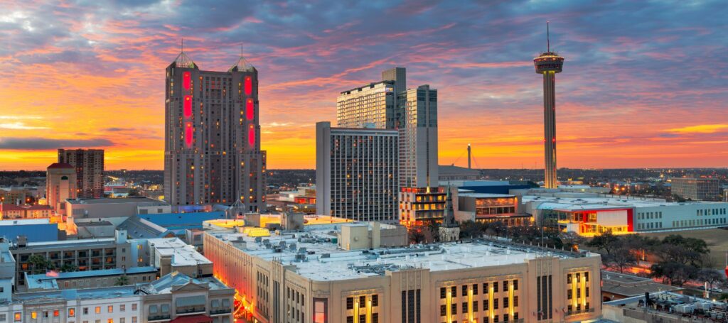 An aerial view of the San Antonio skyline at sunset shows illuminated buildings, including the Tower of the Americas, against a vibrant orange and purple sky