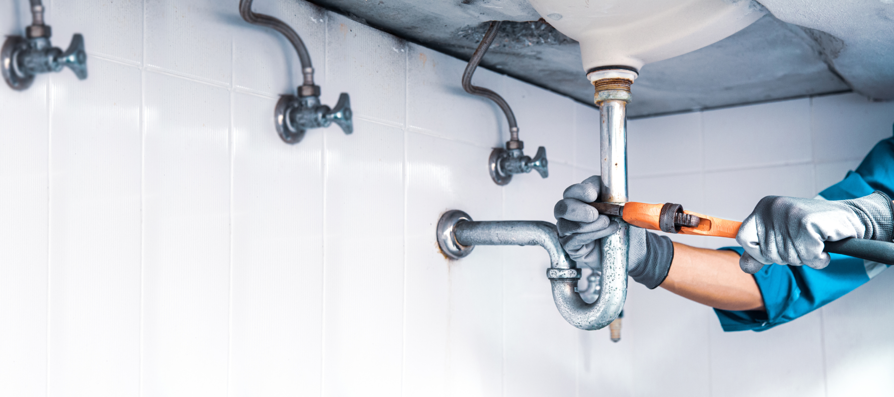 a person wearing grey gloves uses an orange pipe wrench to work on chrome pipes under a sink against a white tiled wall
