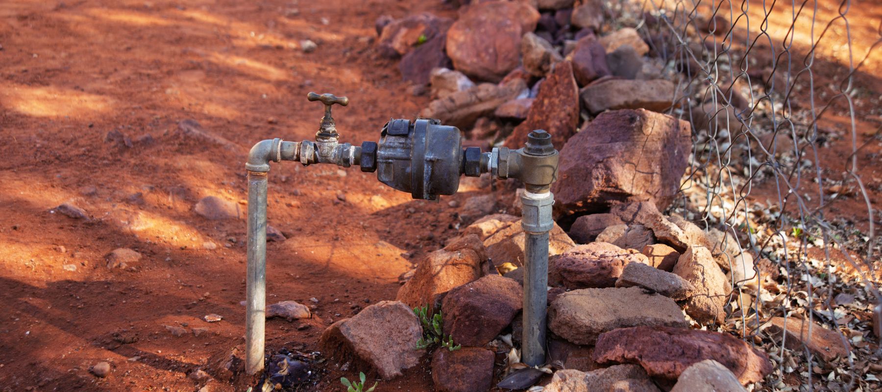 A water meter and pipes are installed in red, rocky soil next to a wire fence