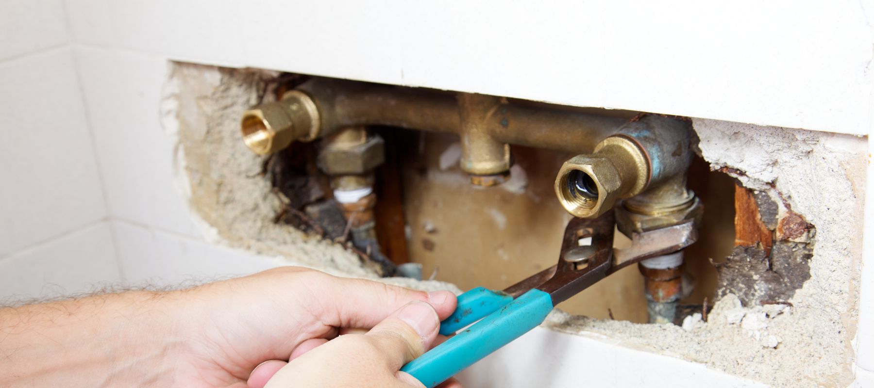 A plumber uses a blue-handled wrench to work on exposed brass pipes embedded in a wall