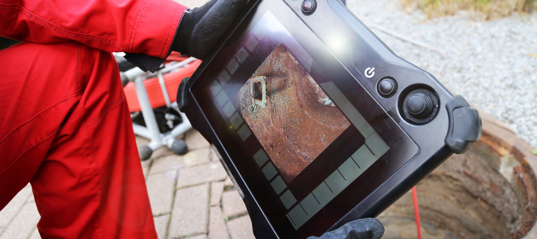 A person wearing gloves holds a device with a screen displaying an image from a pipe inspection camera, with an open manhole in the background