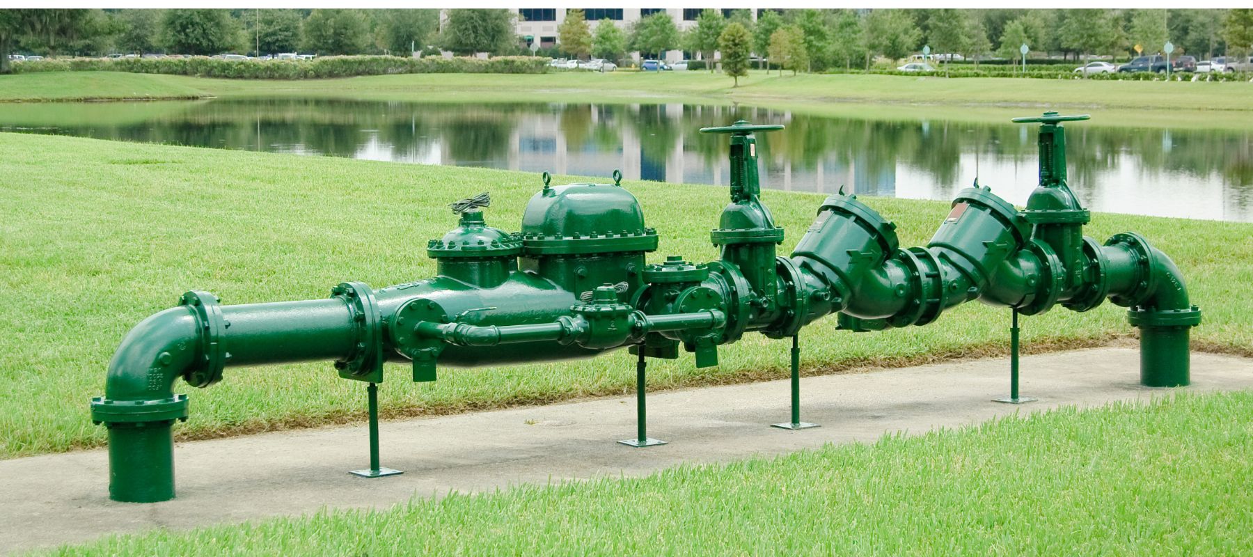 A large green pipe assembly with multiple valves and gauges is displayed horizontally on stands in a grassy area beside a pond, with a building in the background