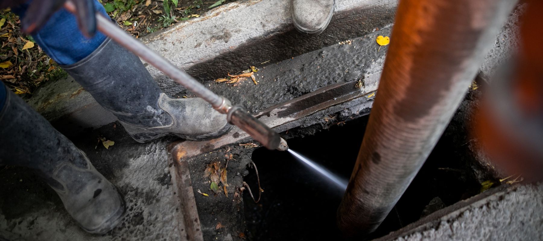 A high-pressure water jet is being used to clear a blockage in a dark manhole, with boots and a large hose visible near the opening