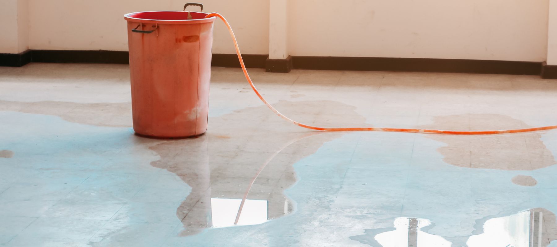 red bucket with an orange hose lies on a wet concrete floor, collecting water from a leak near a window
