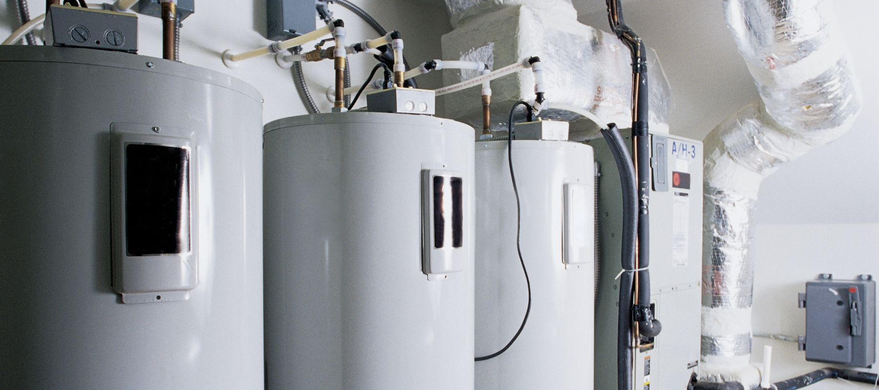 Three large water heaters are lined up in a utility room, connected by various pipes and electrical conduits