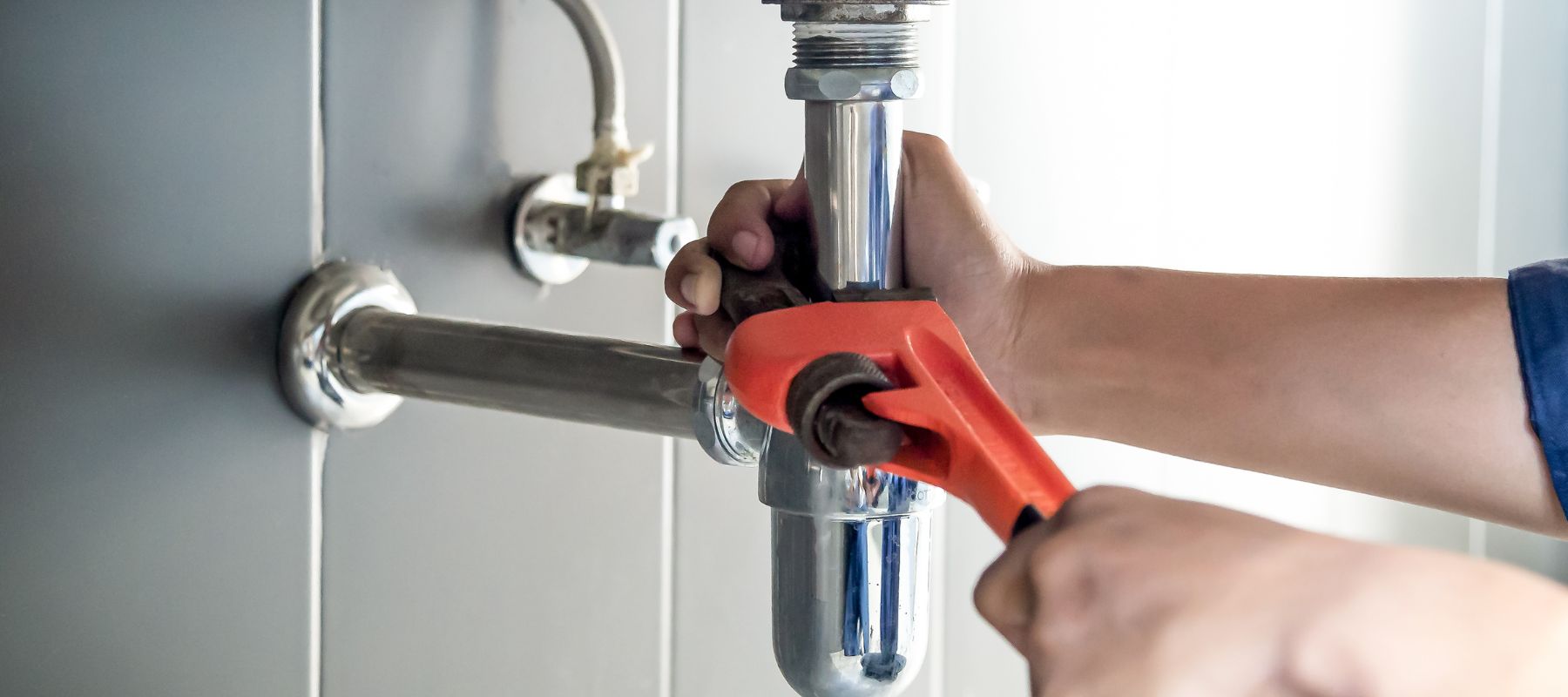 A person's hands use an orange pipe wrench to work on a chrome P-trap pipe under a sink