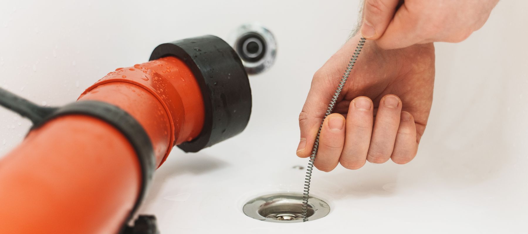 A hand inserts a flexible drain snake into a white bathtub drain, next to a large orange plumbing tool