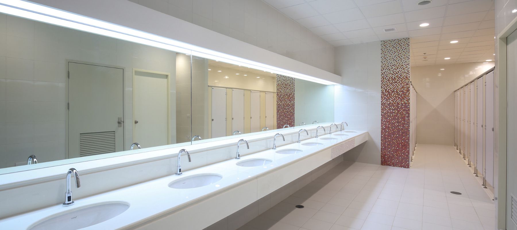 A bright, modern public restroom features a long counter with multiple white sinks and chrome faucets, a large mirror, and individual stalls