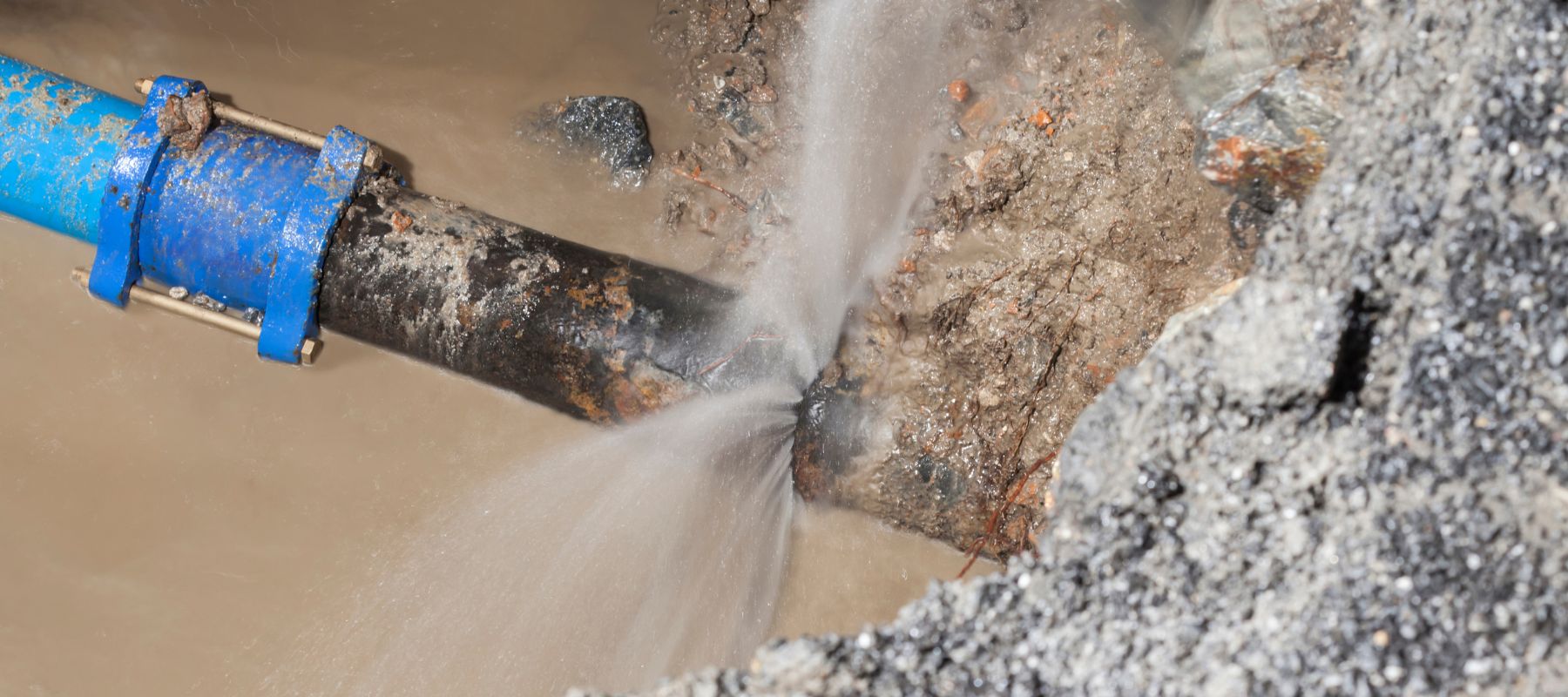 Water fiercely sprays from a burst pipe in a muddy trench, surrounded by gravel and dirt