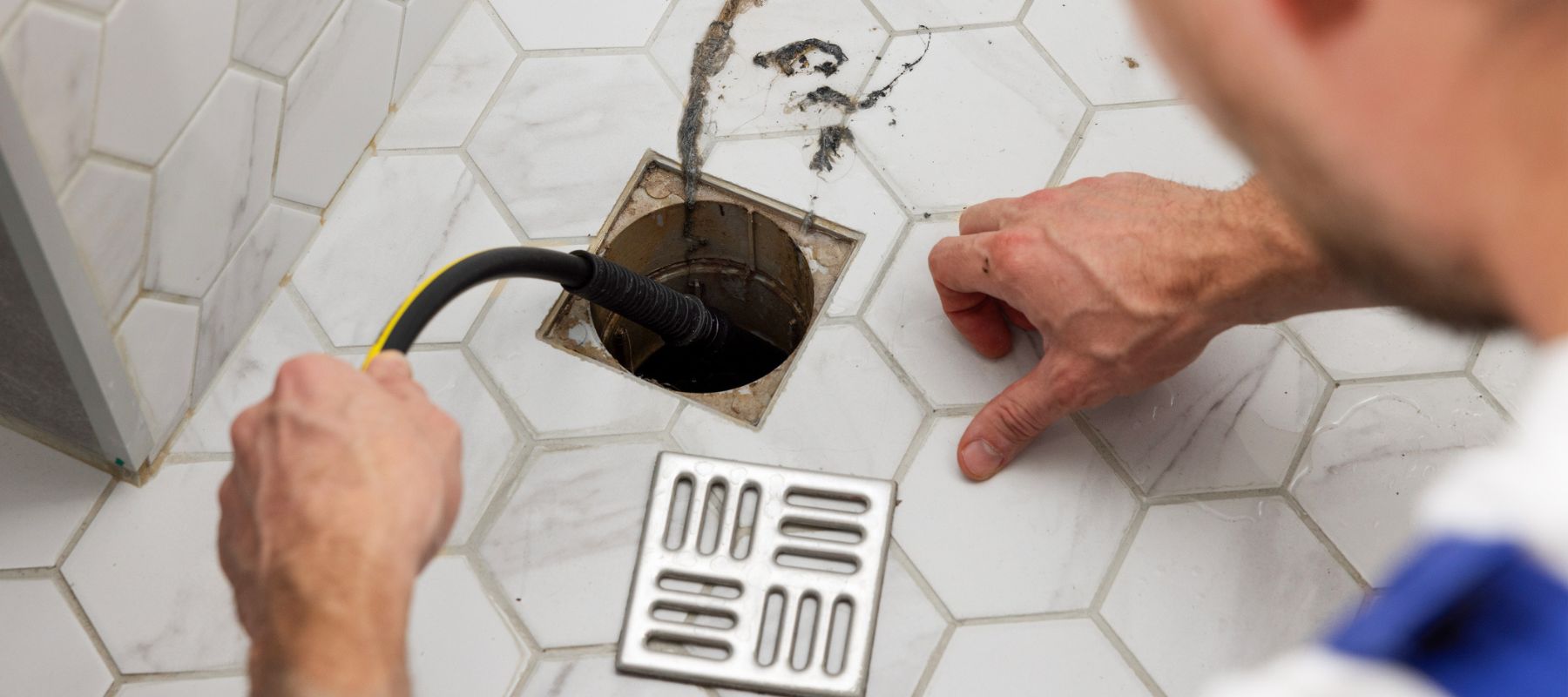 A plumber feeds a black and yellow drain snake into a square floor drain on a white hexagonal tiled floor, with debris visible around the opening