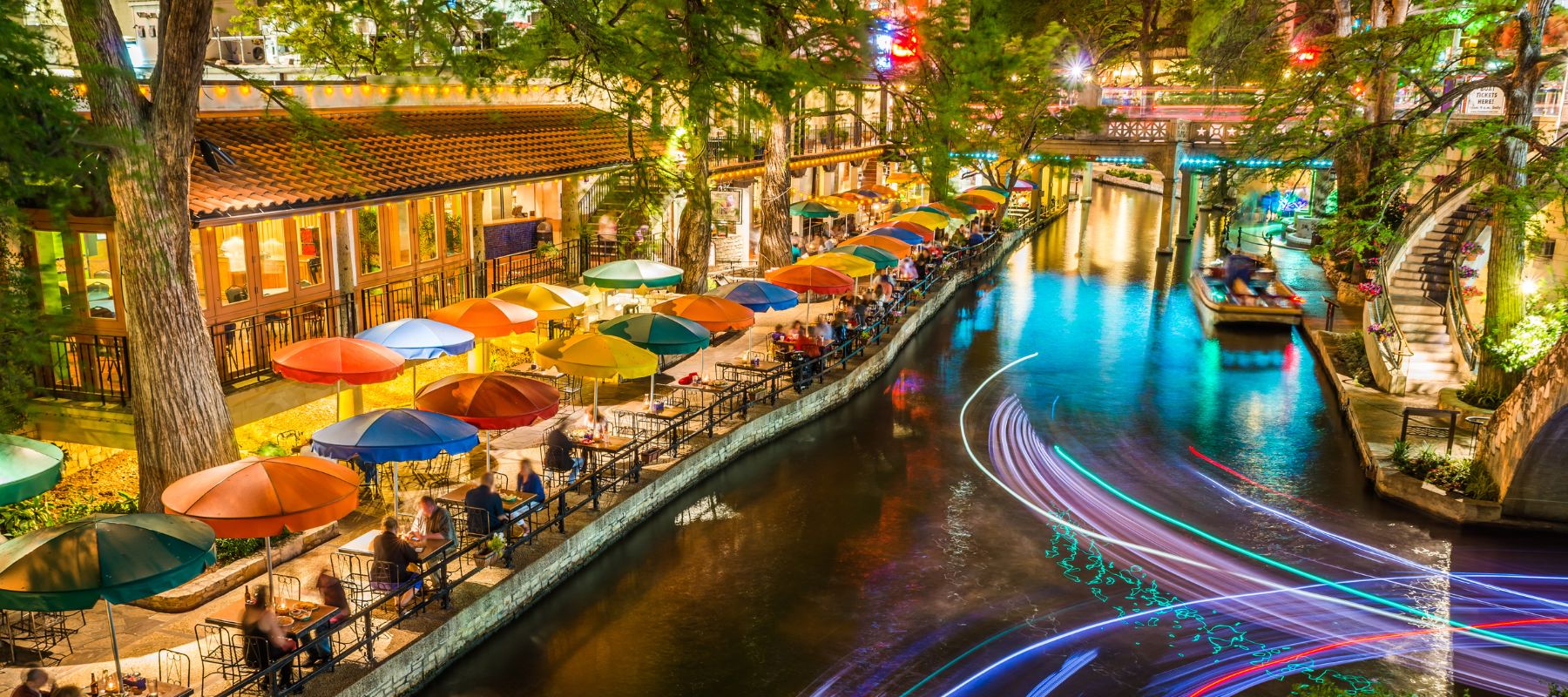 The San Antonio Riverwalk is bustling at night, with restaurants under colorful umbrellas lining the illuminated canal, and light trails from a boat on the water