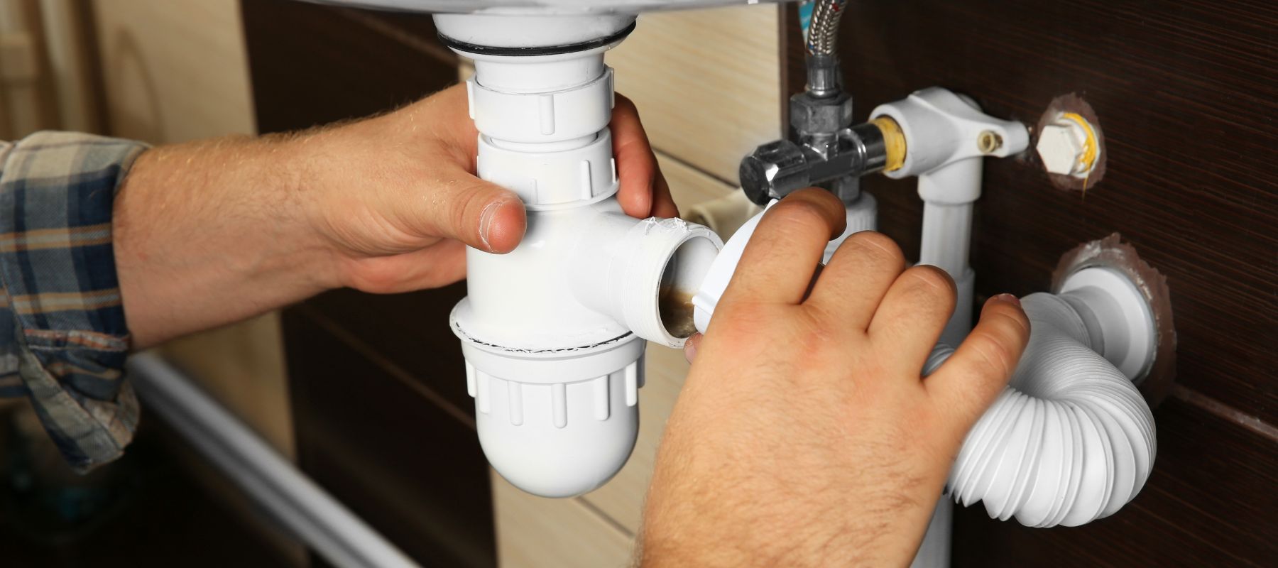 A person's hands are connecting white PVC pipes under a sink, with various plumbing fixtures visible behind