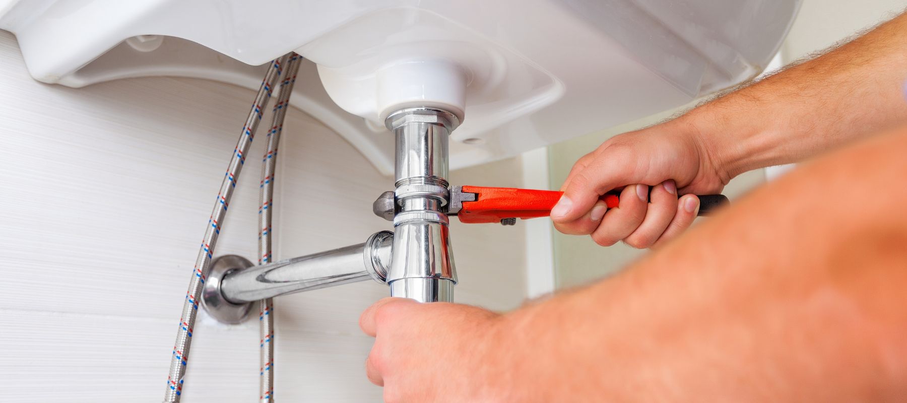 A plumber uses a red pipe wrench to tighten a chrome pipe connection under a white sink