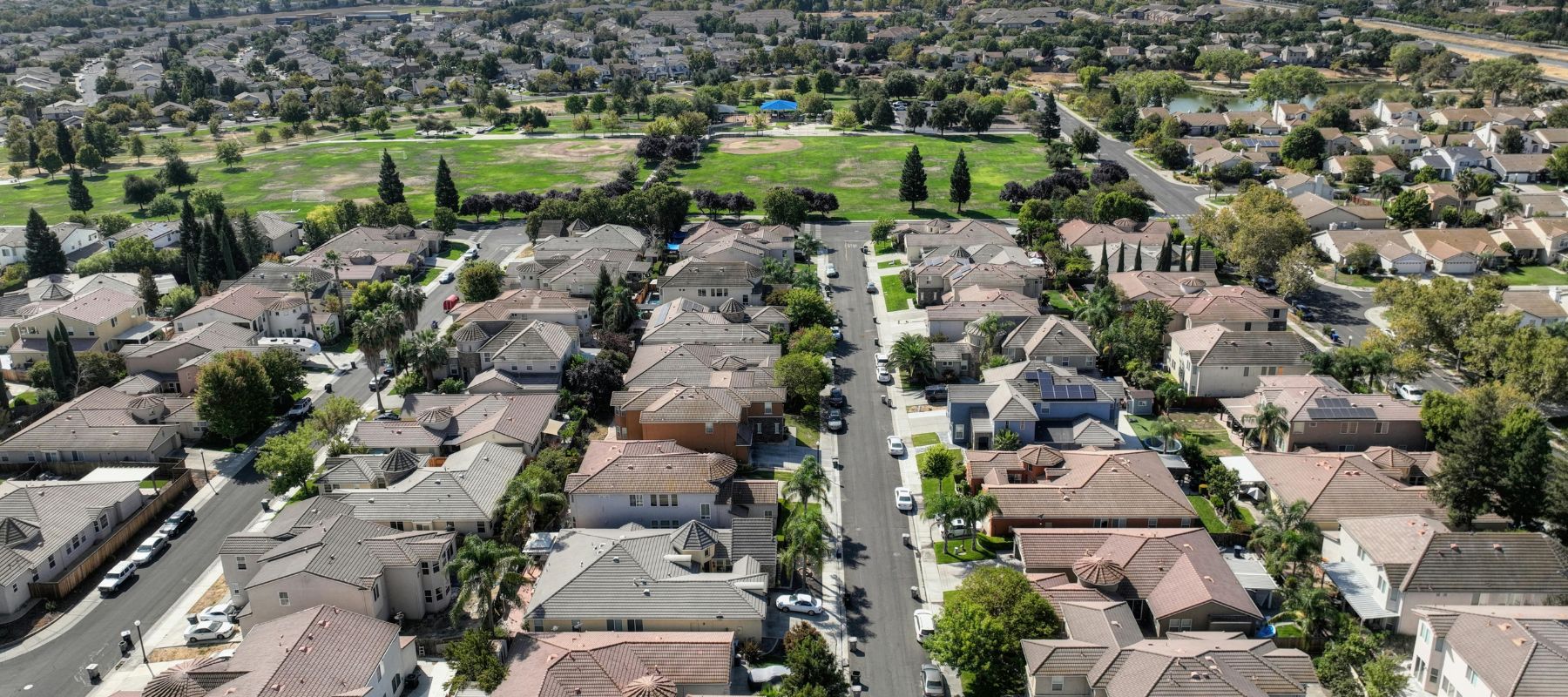 An aerial view captures a suburban neighborhood with numerous houses and green spaces, including a park in the distance