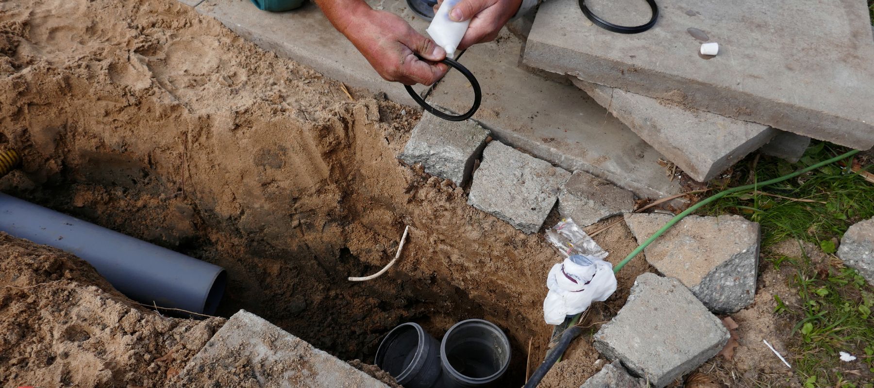 A person works in a trench, holding plumbing supplies near buried pipes and loose dirt
