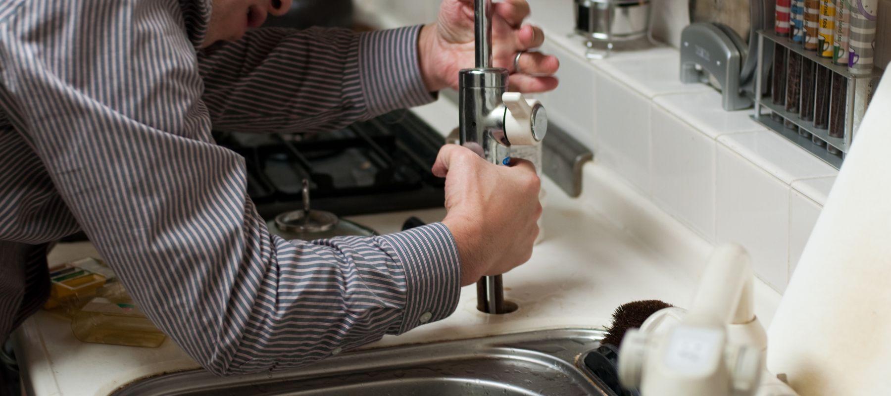 A person's hands are actively working on the base of a kitchen faucet, appearing to install or repair it above a sink
