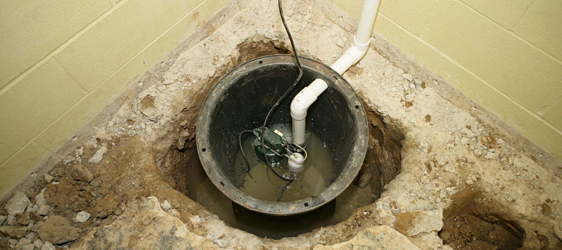 A sump pump is visible in a round pit dug into the ground, surrounded by dirt and concrete