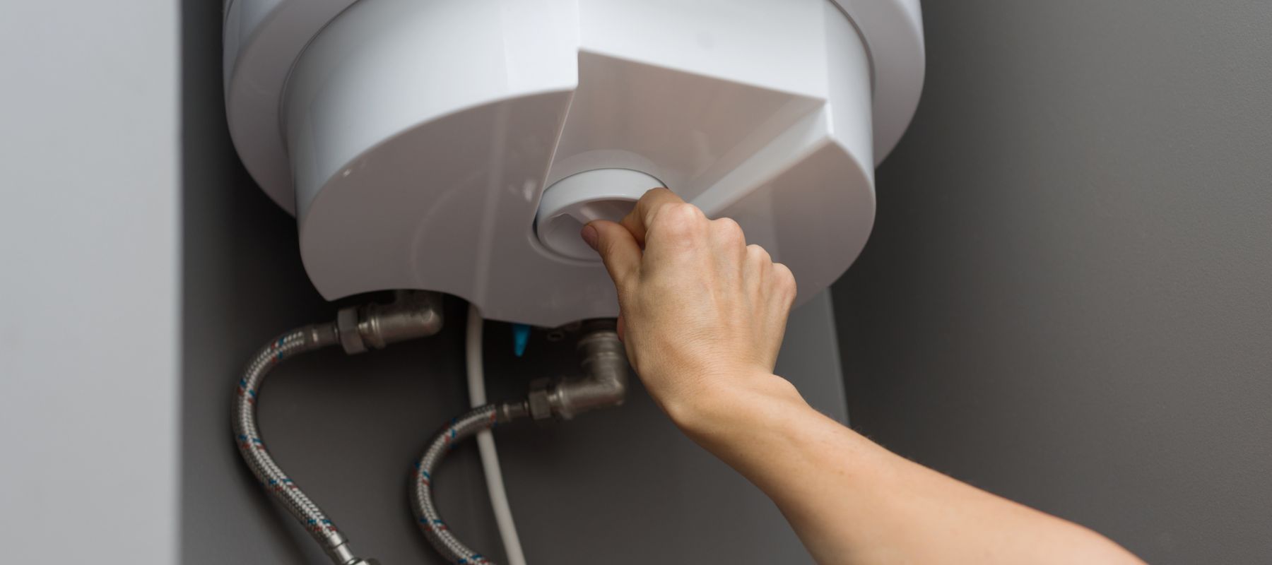 A hand adjusts a white dial on the bottom of a white water heater, with pipes and an electrical outlet visible below