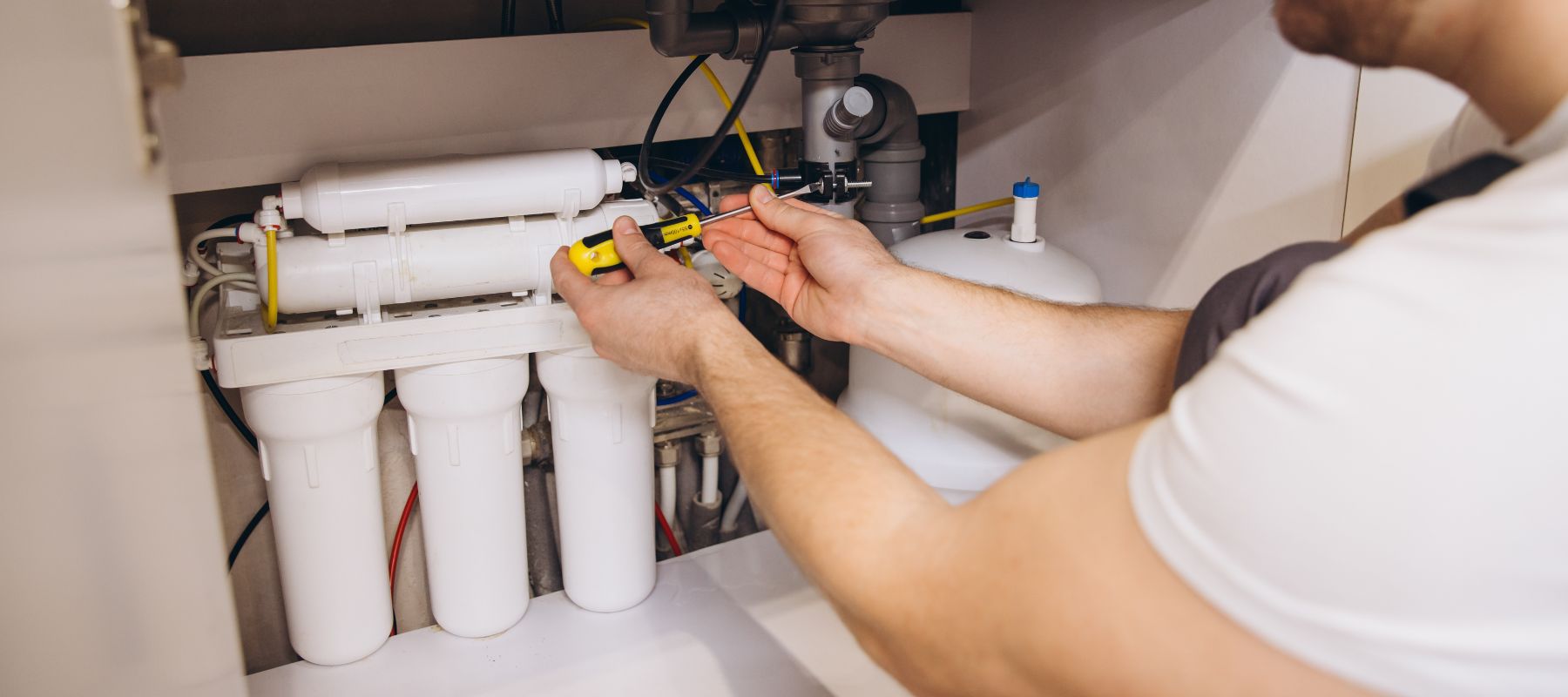 A person uses a screwdriver to work on a white multi-stage water filtration system installed under a sink