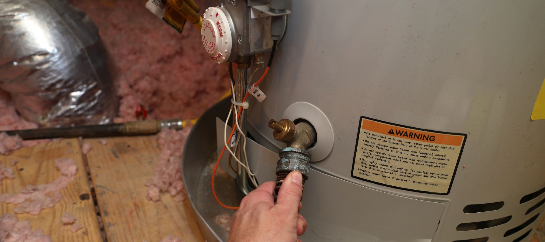 A person's hand disconnects a pipe from a water heater with an "Warning" label, surrounded by insulation