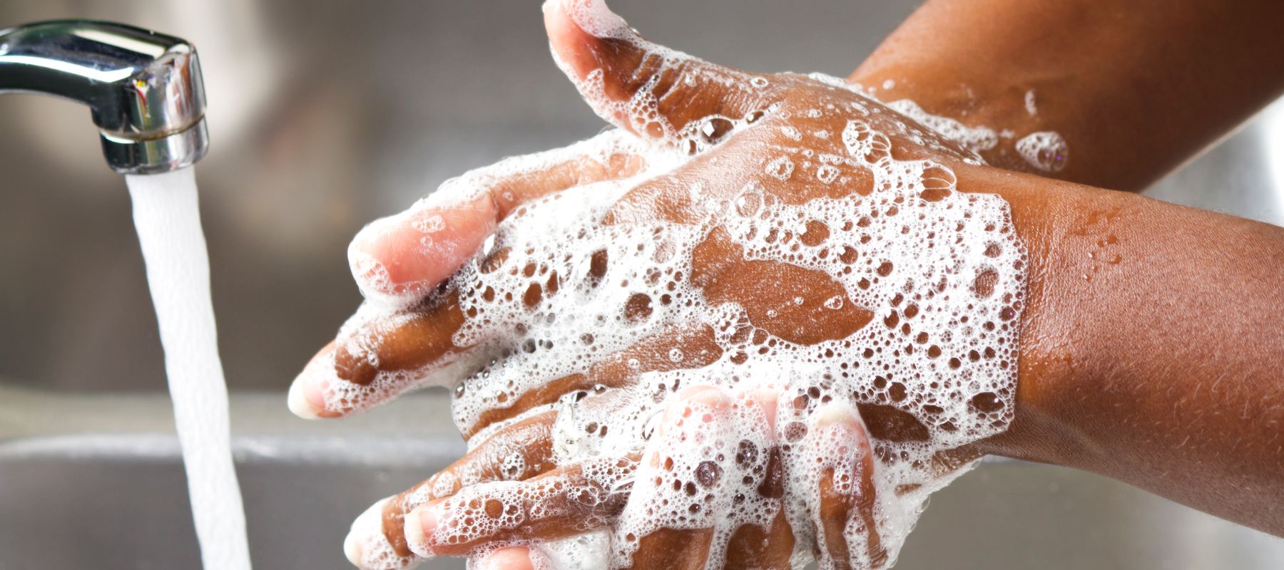 Soapy hands are being rinsed under a stream of water from a chrome faucet in a stainless steel sink