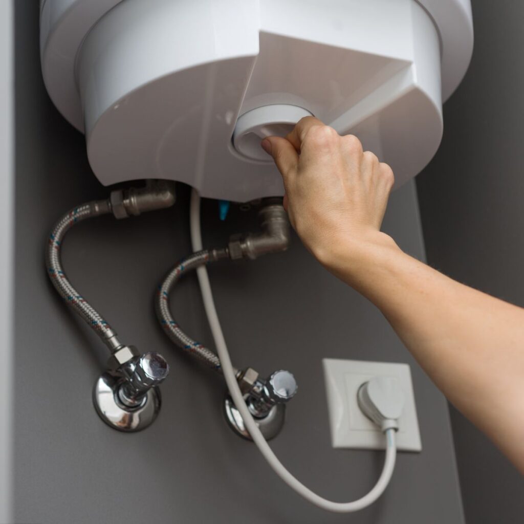 A hand adjusts a white dial on the bottom of a white water heater, with pipes and an electrical outlet visible below.