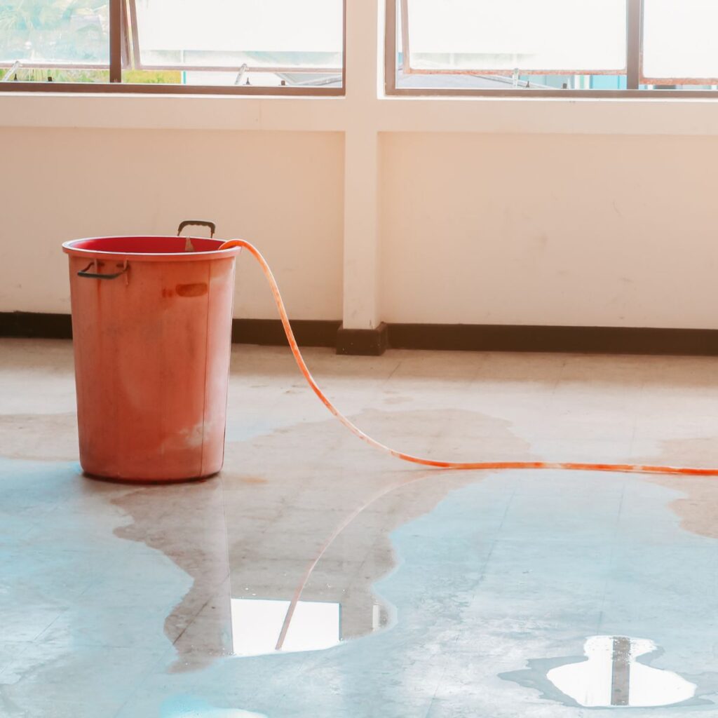 A red bucket with an orange hose lies on a wet concrete floor, collecting water from a leak near a window