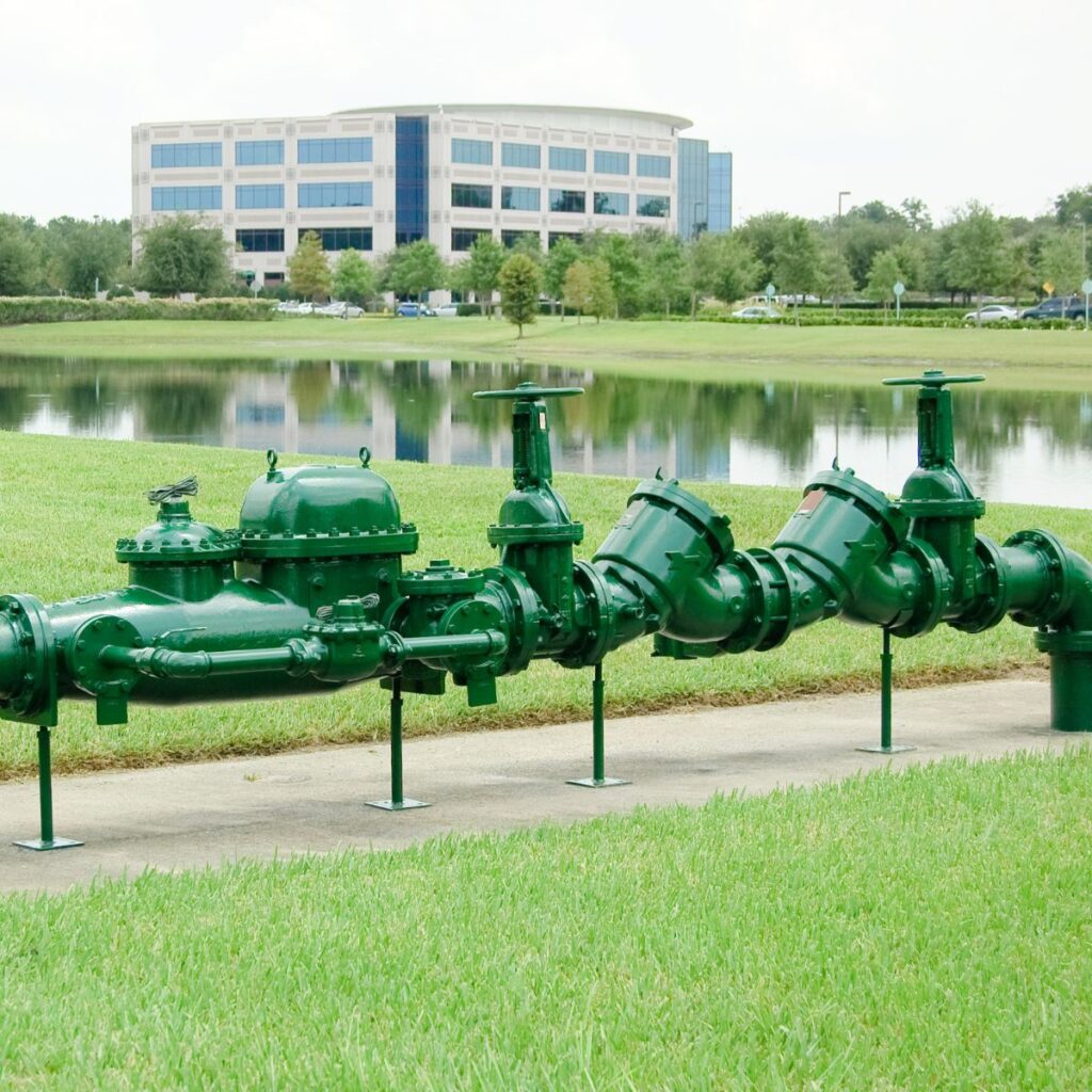 A large green pipe assembly with multiple valves and gauges is displayed horizontally on stands in a grassy area beside a pond, with a building in the background