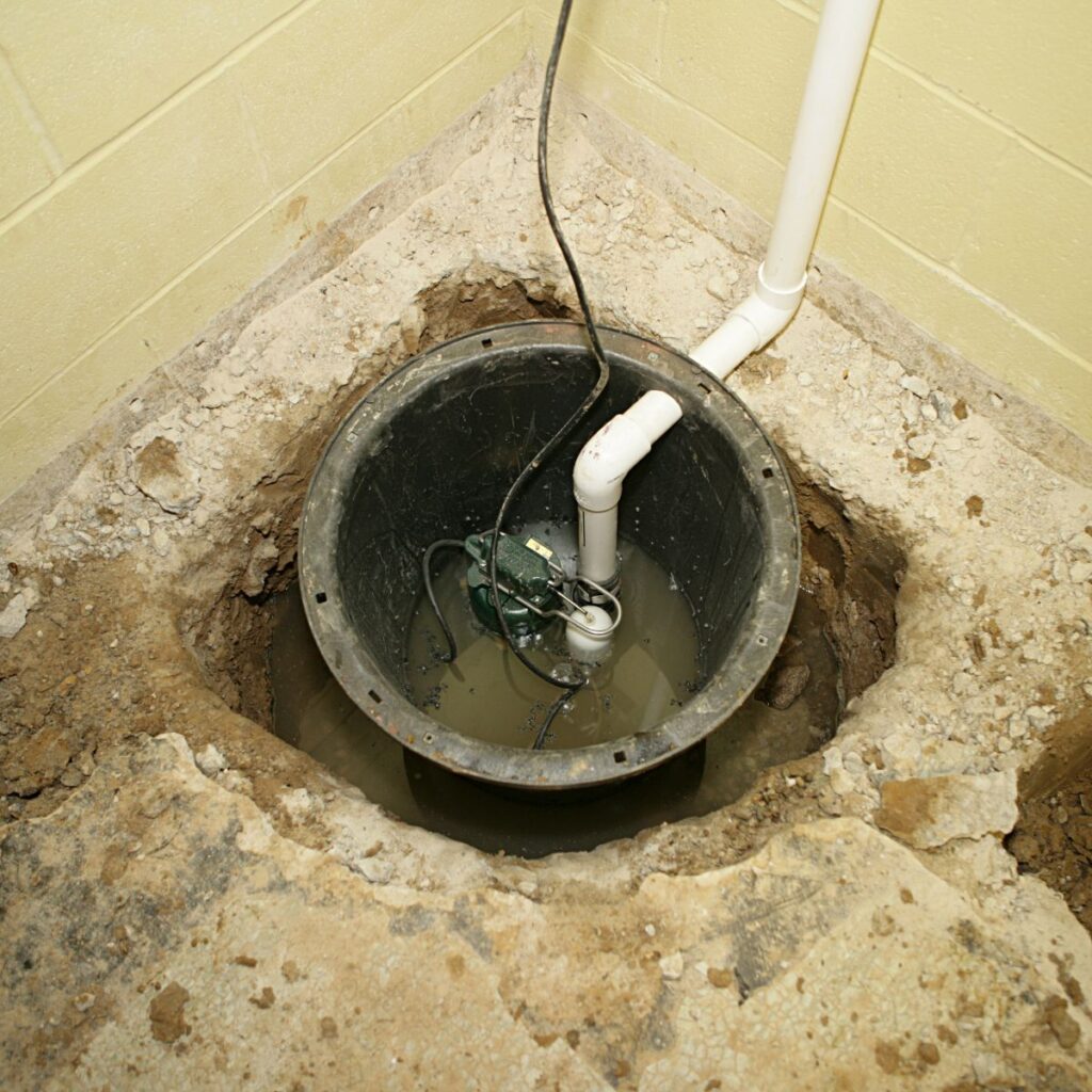 A sump pump is visible in a round pit dug into the ground, surrounded by dirt and concrete