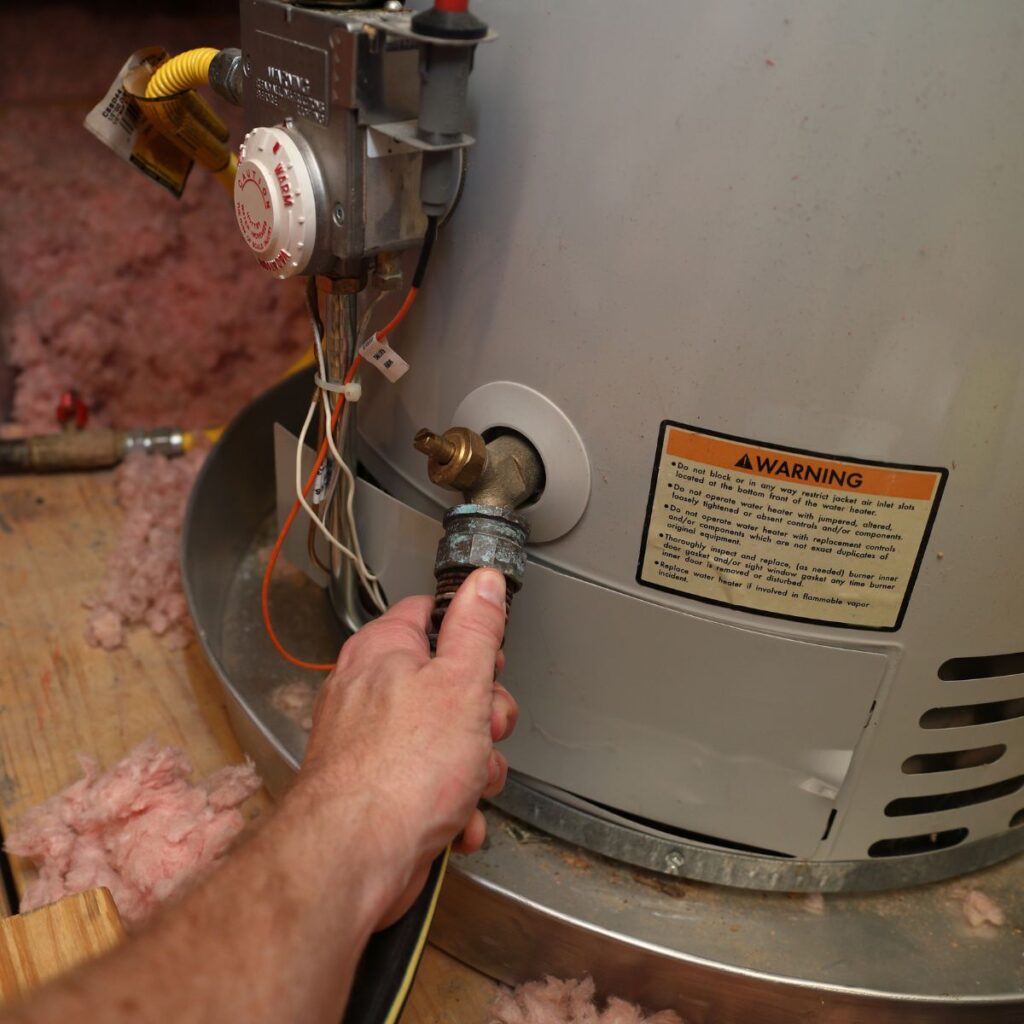 A person's hand disconnects a pipe from a water heater with an "Warning" label, surrounded by insulation