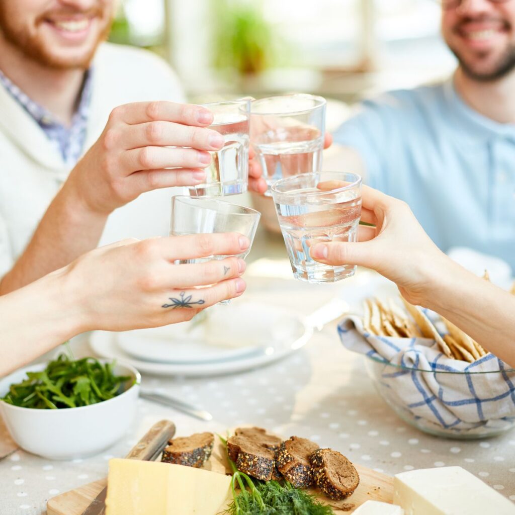 Several people are making a toast with small glasses of water at a table laden with cheese, bread, and a green salad