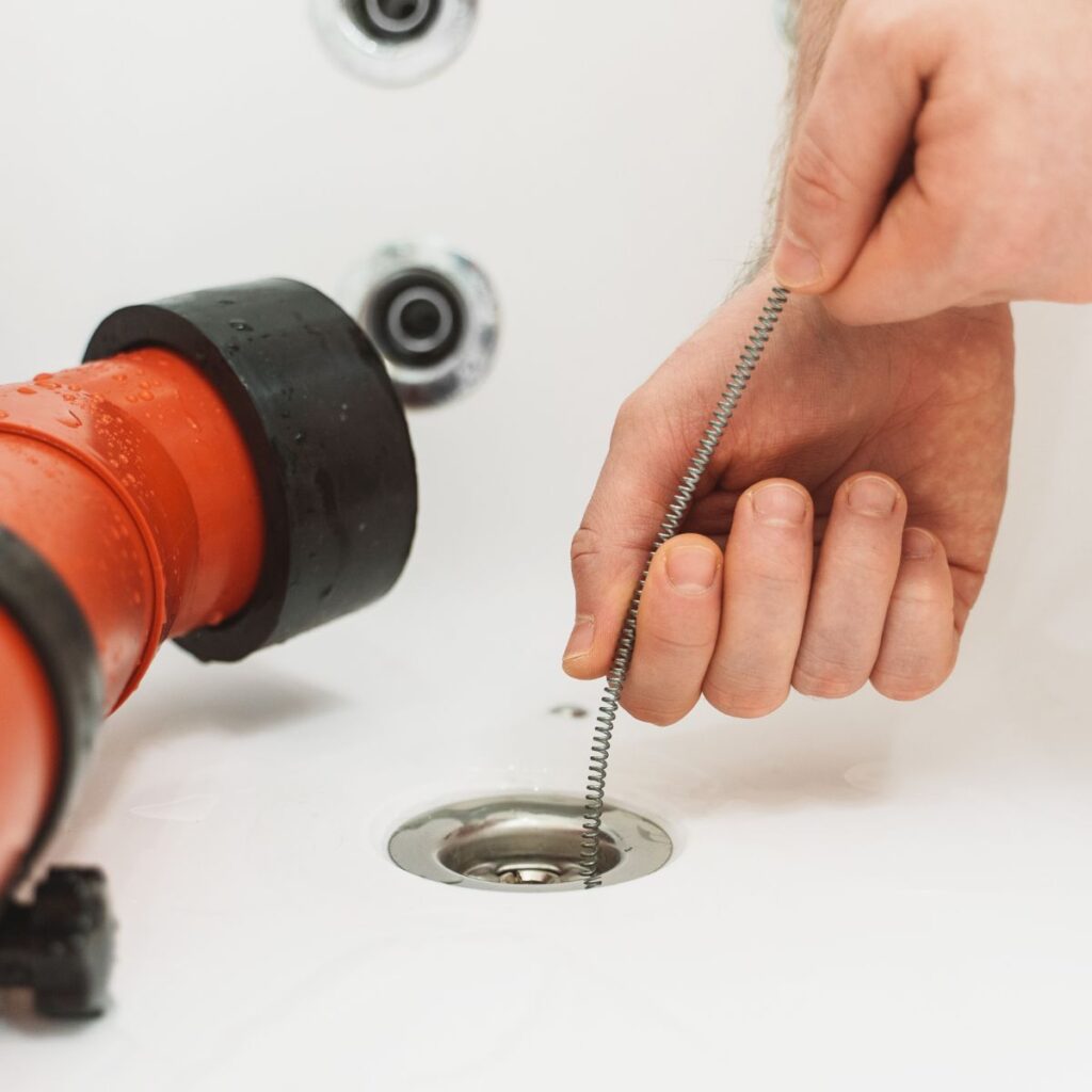 A hand inserts a flexible drain snake into a white bathtub drain, next to a large orange plumbing tool