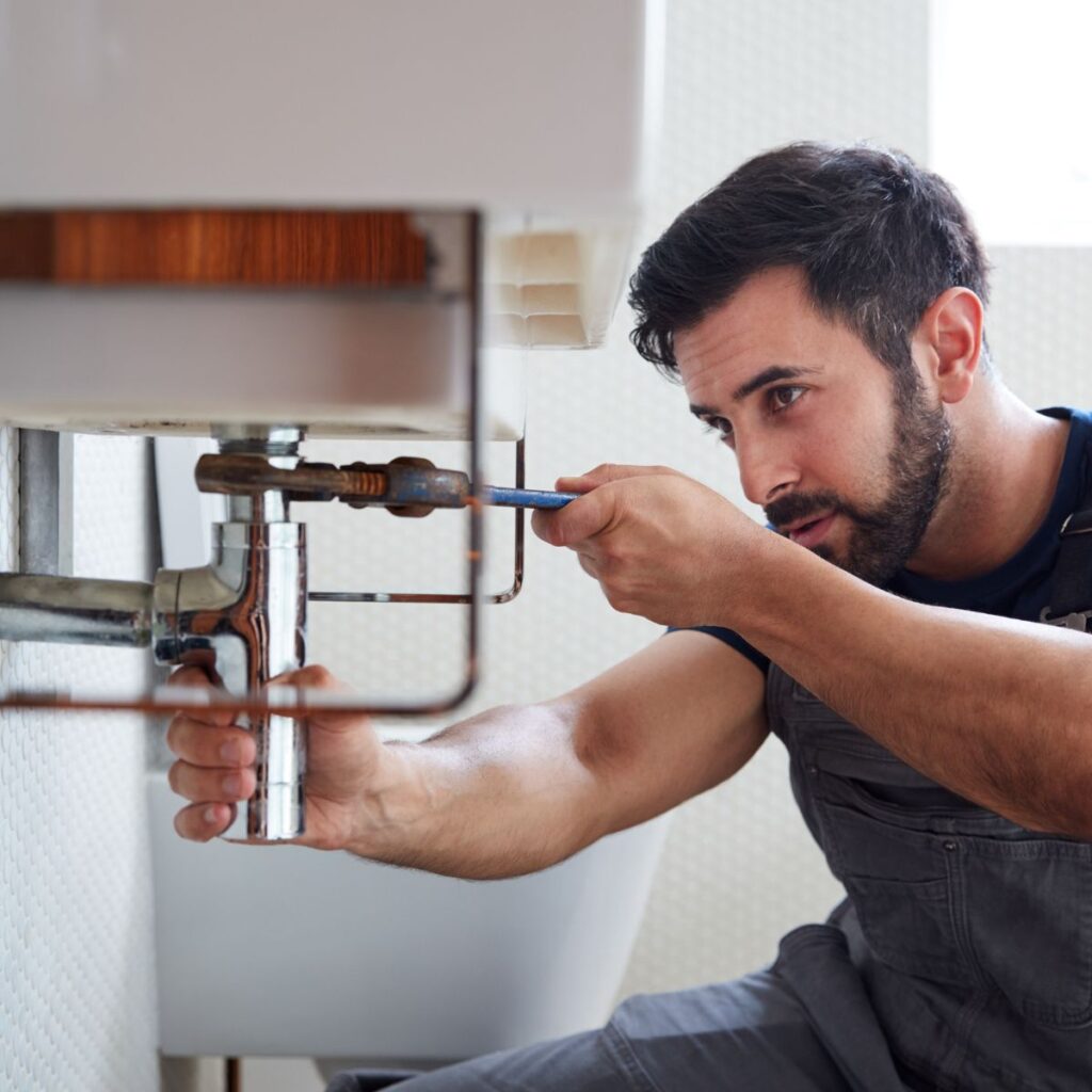 A plumber with a beard uses a small wrench to work on chrome pipes under a bathroom sink