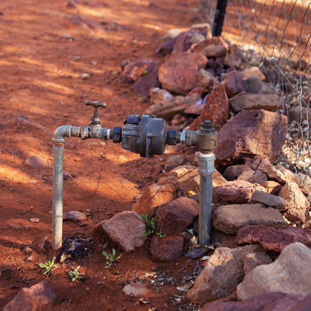 A water meter and pipes are installed in red, rocky soil next to a wire fence