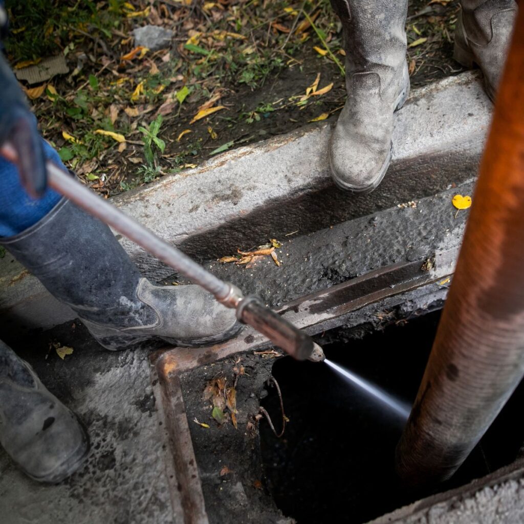 A person's booted feet stand near an open manhole, using a high-pressure hose to clear debris from the drain, with a large orange hose also entering the opening