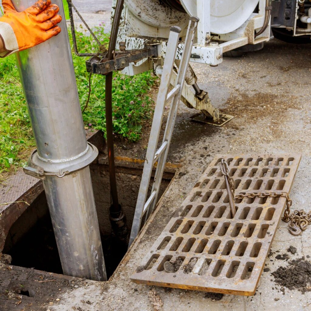 A large metal pipe is being lowered into an open manhole next to a ladder and a utility truck, with a metal grate on the ground