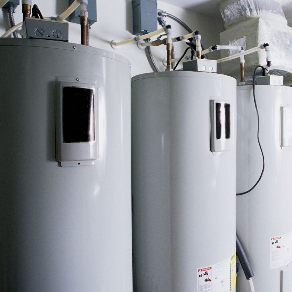 Three large water heaters are lined up in a utility room, connected by various pipes and electrical conduits