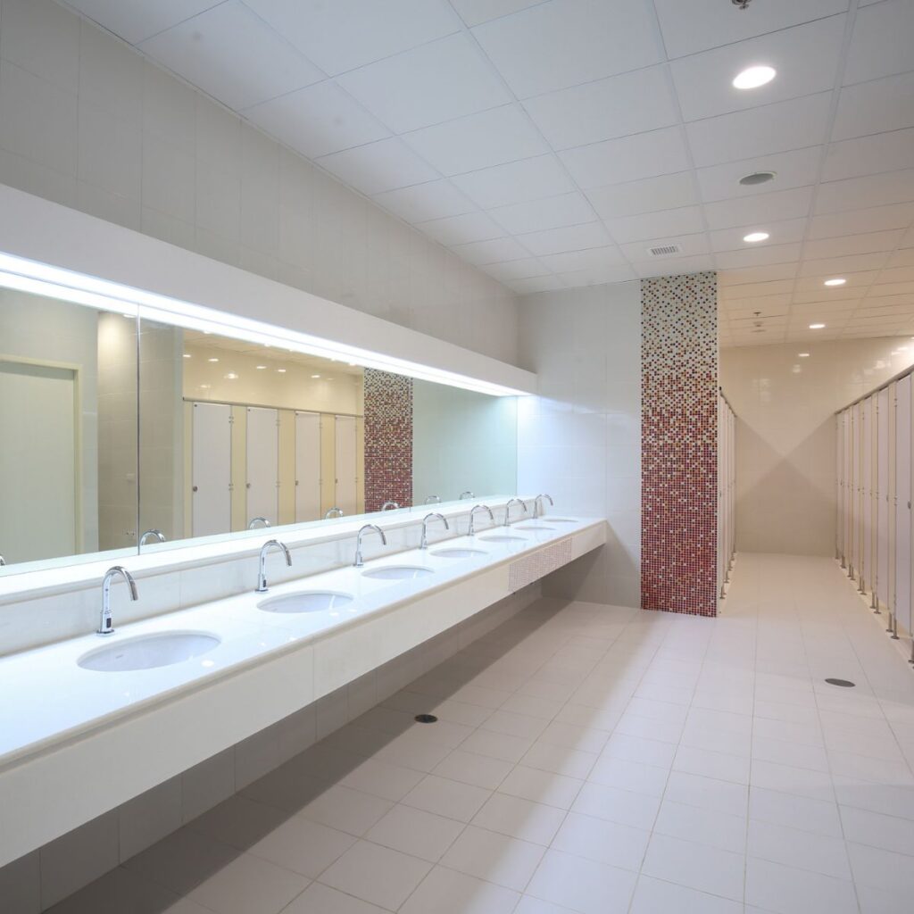 A bright, modern public restroom features a long counter with multiple white sinks and chrome faucets, a large mirror, and individual stalls