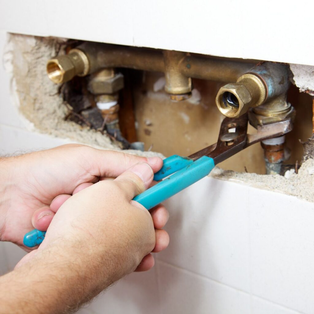 A plumber uses a blue-handled wrench to work on exposed brass pipes embedded in a wall.